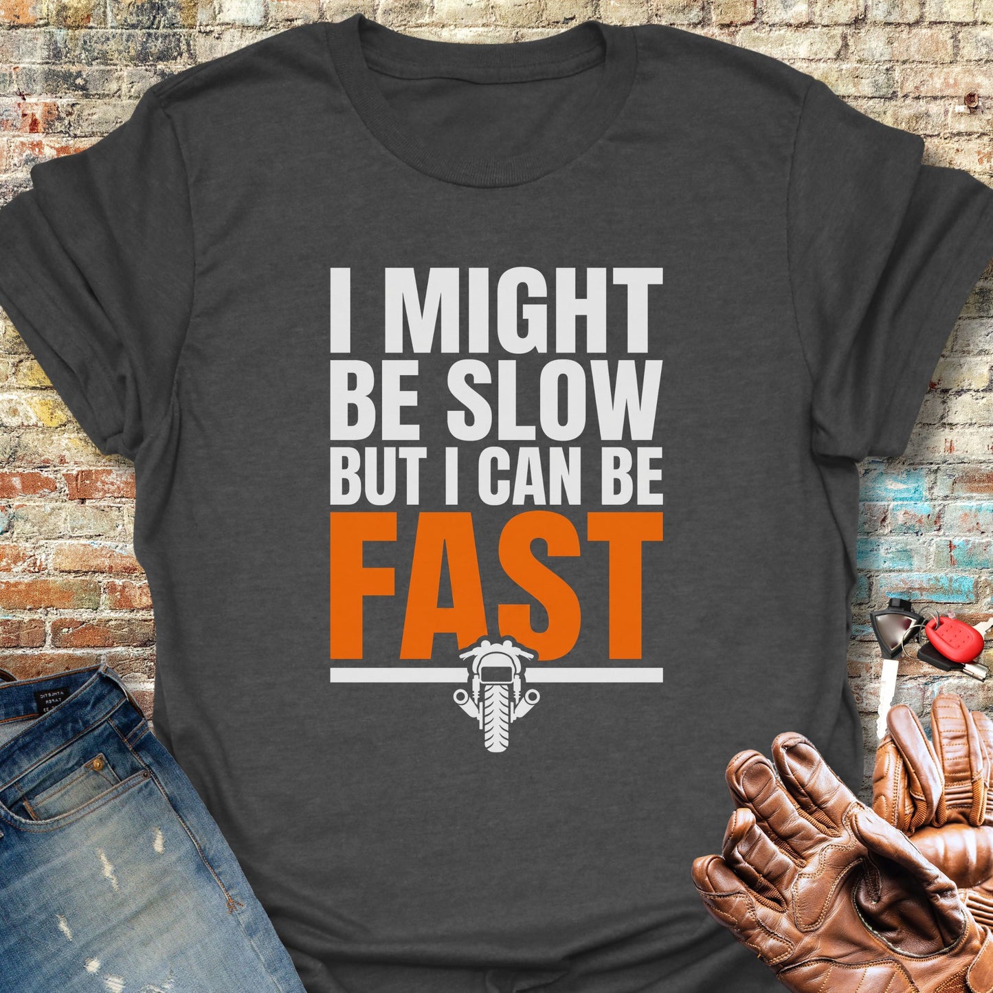 A dark charcoal gray graphic t-shirt featuring the text 'I MIGHT BE SLOW BUT I CAN BE FAST' in white and orange lettering, with a small motorcycle graphic underneath. The shirt is styled in a flat-lay arrangement with a brick wall background, blue jeans, and a brown leather baseball glove.